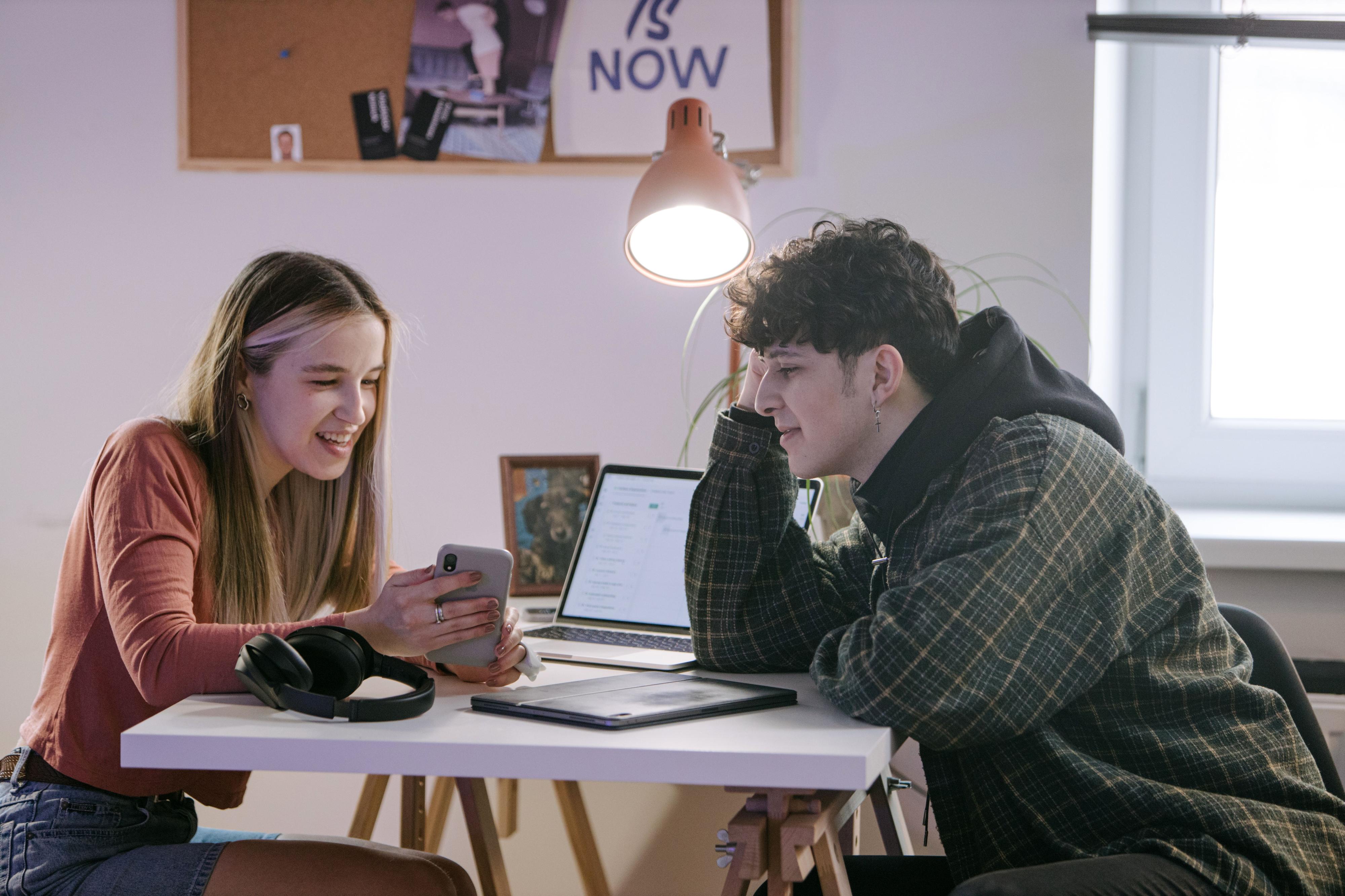 Two students chatting while using a smartphone in a shared indoor space. 